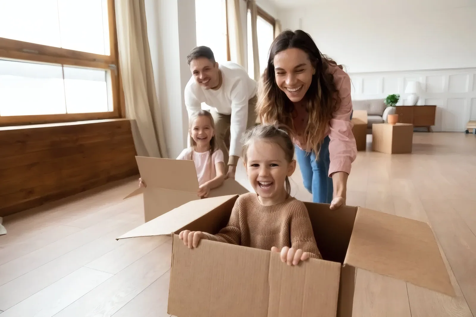 Una familia alegre jugando en un salón luminoso y soleado, los padres empujando por el suelo a sus hijas que ríen dentro de cajas de cartón.